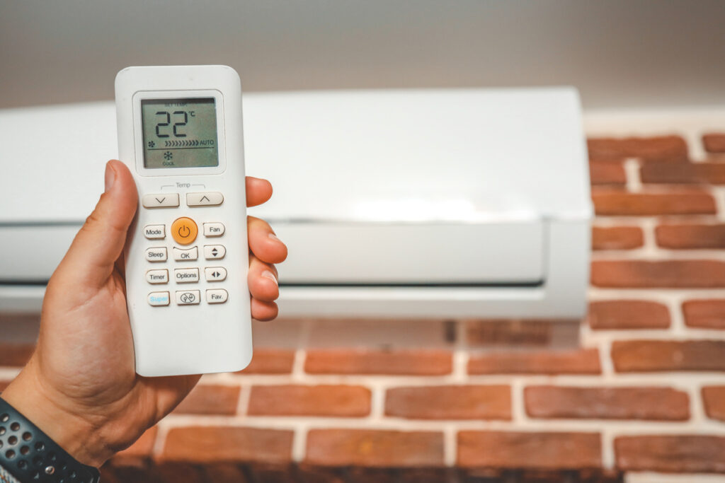 pessoa branca segurando com uma mao o controle de ar condicionado e ao fundo em um uma parede de tijolos o ar condicionado na parede.