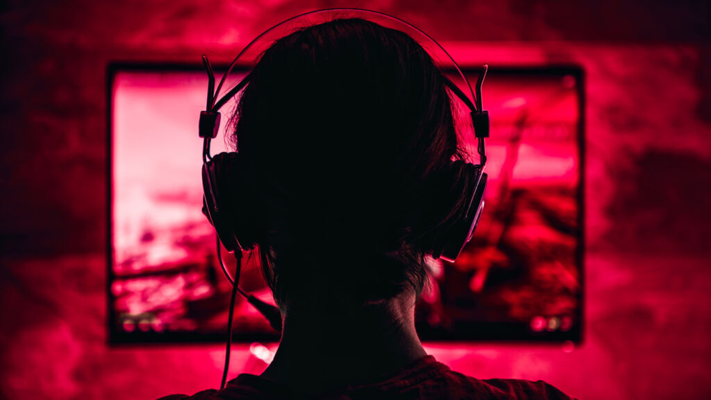 Pessoa de costas usando headset de áudio diante de um monitor em um ambiente com iluminação vermelha, representando gaming ou streaming de videogame.