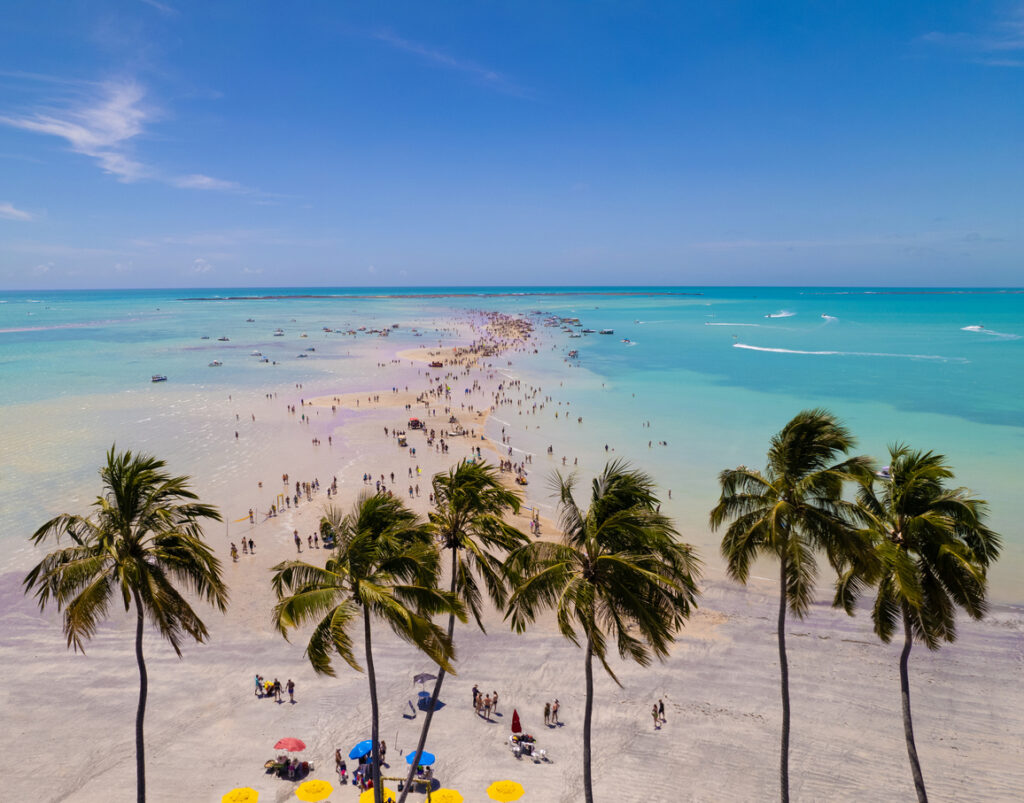 Praia paradisíaca com águas cristalinas, areia branca e palmeiras ao pôr do sol, repleta de turistas desfrutando do litoral.