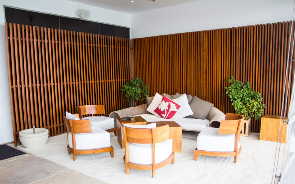 Sala de estar moderna com sofá bege, cadeiras brancas com estrutura de madeira, tapete branco, plantas naturais e painel de madeira decorativa ao fundo, criando ambiente acolhedor e elegante.